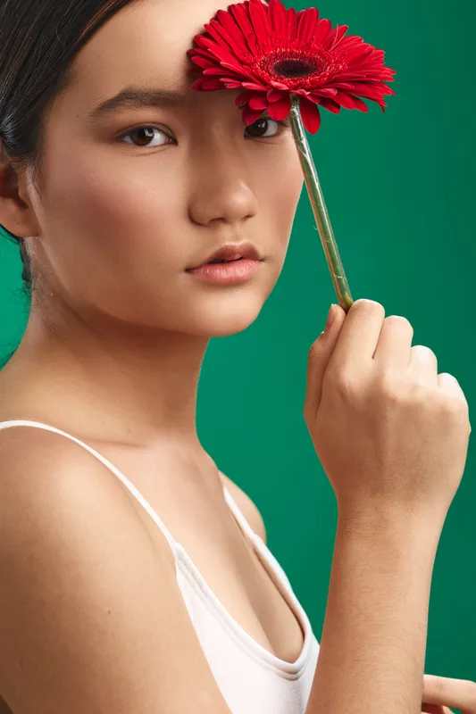 Asian woman holds a vibrant red flower close to her face, gazing softly at the camera in a studio setting with a lush green background.