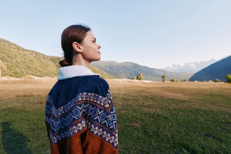 Woman portrait in sweater profile standing in a meadow with mountains and valley in background, outdoors nature scene showing calm horizon, serene expression and scenic landscape.