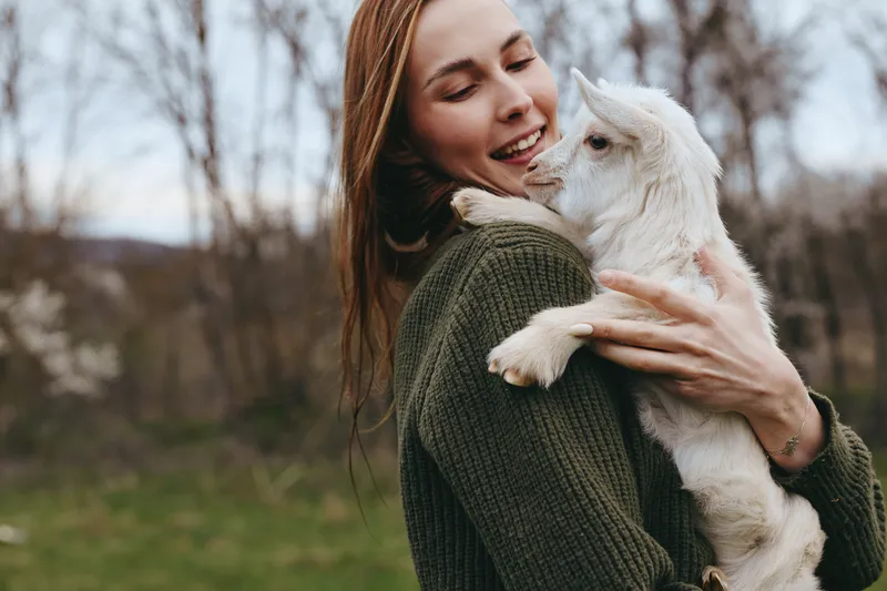 Joyful outdoor moment, a woman hugs a white goat kid, smiling with closed eyes, soft breeze, earthy tones, cozy sweater, rustic background, lifestyle scene