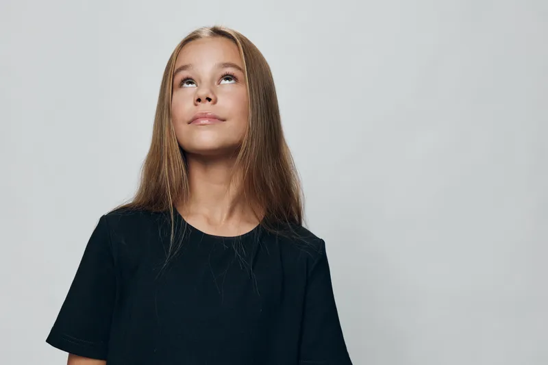 Girl with long hair looking up against light background with thoughtful expression wearing simple black shirt in studio portrait with neutral mood.