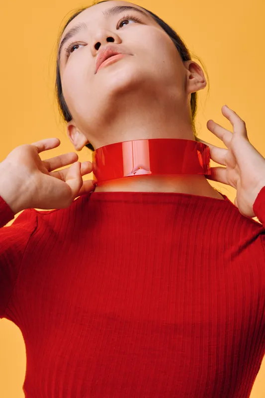 Woman with red ribbon around neck in red top on yellow background, hands touching neck, closeup portrait with minimal style and bright color contrast in studio light.