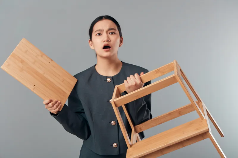 Woman holding broken wooden chair and seat with surprised expression standing against grey background indoors. Female with damaged furniture, repair concept and home interior scene.