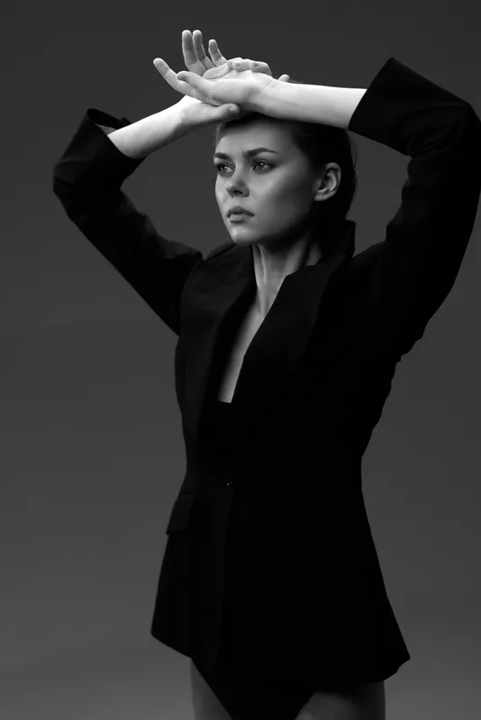 Black and white portrait of a serious woman in a black jacket with raised arms, expressing thoughtful and confident emotions.