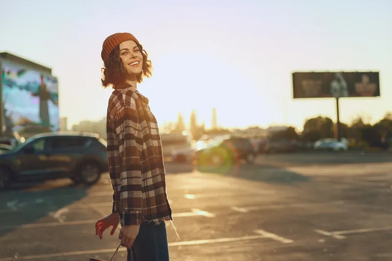 Woman smile beanie urban sunset candid lifestyle golden authenticity mindful living emotional storytelling in a parking lot backlit at golden hour with relaxed joyful expression and city warmth