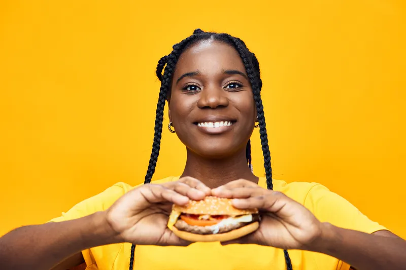 Happy young African woman holding a delicious hamburger against a vibrant yellow background, showcasing a joyful expression and a casual summer style