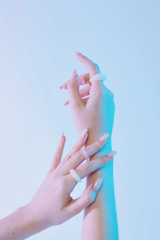 Close-up of female hands with long nails and translucent rings on fingers. Soft blue lighting highlights beauty and fashion details on a smooth skin background. Concept for style.
