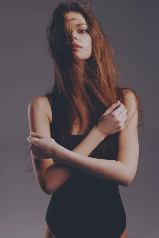 Young woman with long hair posing in black outfit with serious expression against grey background.