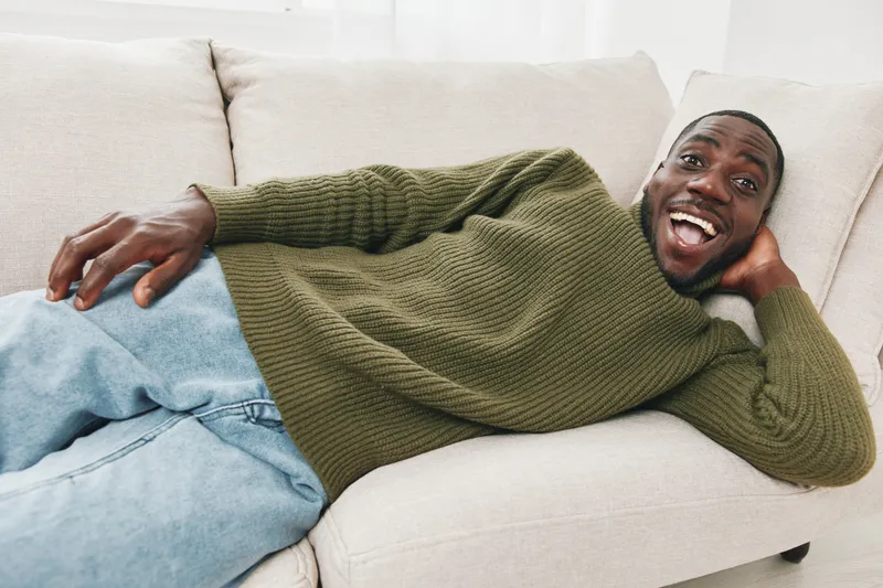 Happy African man relaxing on a light sofa in casual sweater and denim jeans, conveying joy and comfort in a cozy home setting for lifestyle imagery