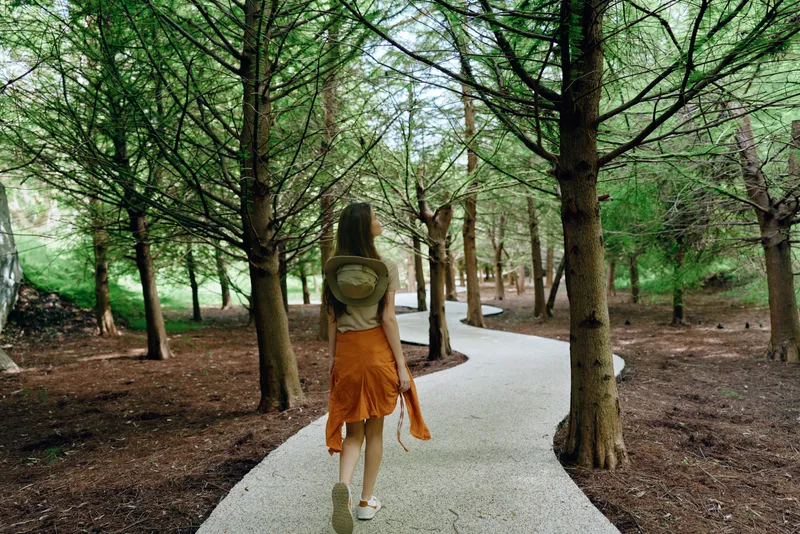 Woman walking forest path with backpack and hat among tall trees and nature, curved gravel walkway, casual skirt and daylight creating a serene travel scene for outdoor exploration.