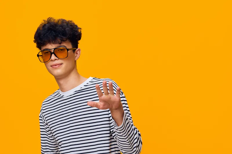 young man in striped shirt wearing sunglasses posing with raised hand against bright orange background, casual style and confident expression