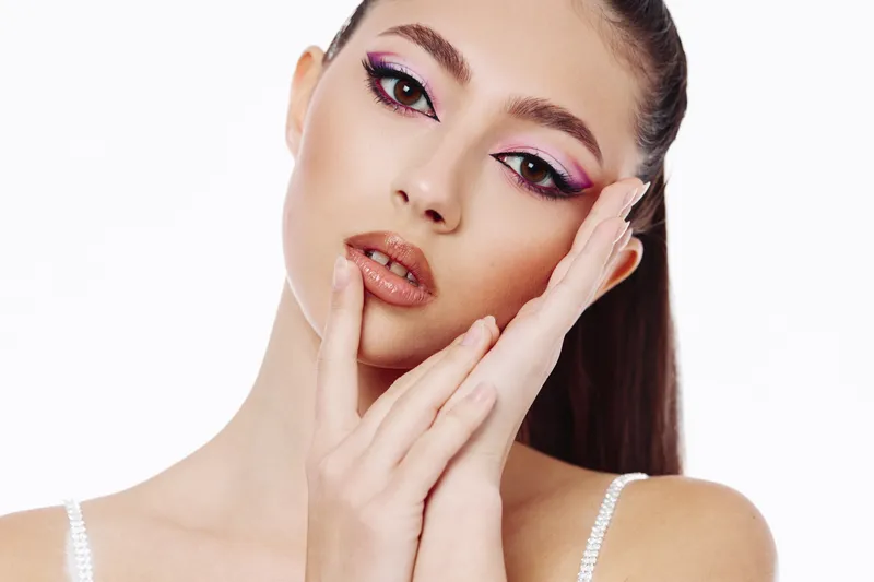 Young woman with creative colorful makeup and glossy lips poses with hand near face on white background. Focus on beauty, fashion, and skincare elegance. Studio lighting highlights details.