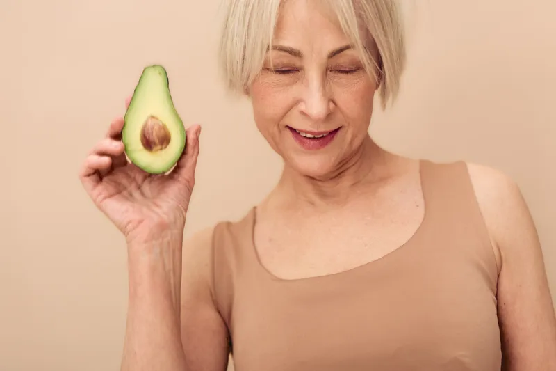 Smiling mature woman with short hair holds half avocado close to face on beige background. Concept of healthy eating, natural lifestyle, nutrition, and wellness for older adults.