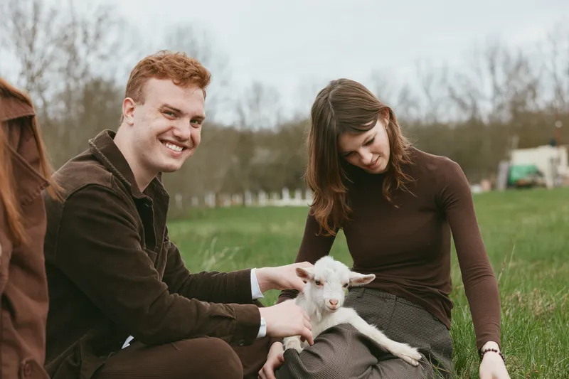 Young couple enjoys spending time outdoors with a baby goat in a grassy field while smiling and interacting in a relaxed lifestyle moment.