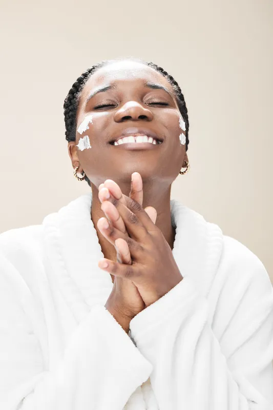 Smiling African woman applying skincare cream, wearing a white bathrobe, with a relaxed ambiance and soft neutral background Focus on skincare in wellness routine