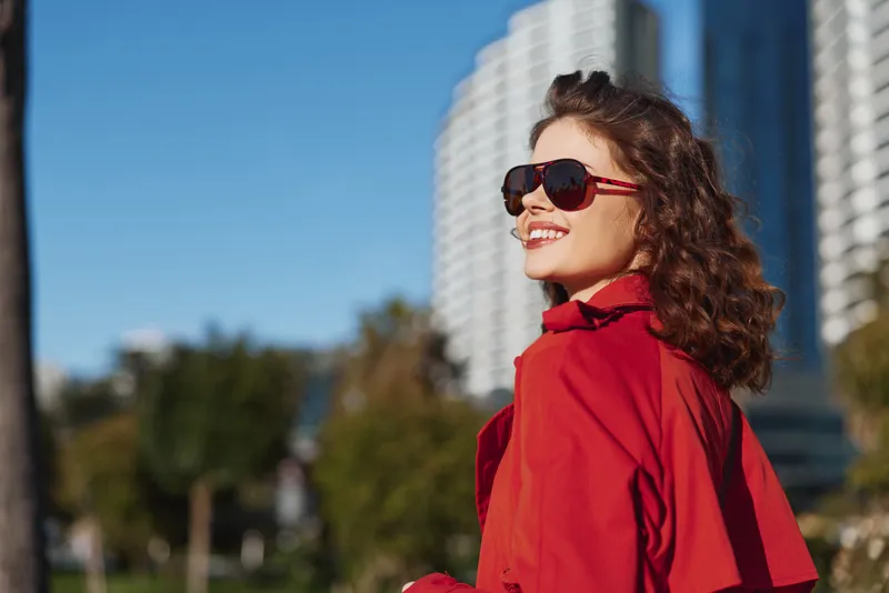 Stylish woman wearing a bright red jacket smiles happily in sunny city weather, posing confidently beside modern high rise buildings under a clear blue sky, creating a vibrant streetstyle moment