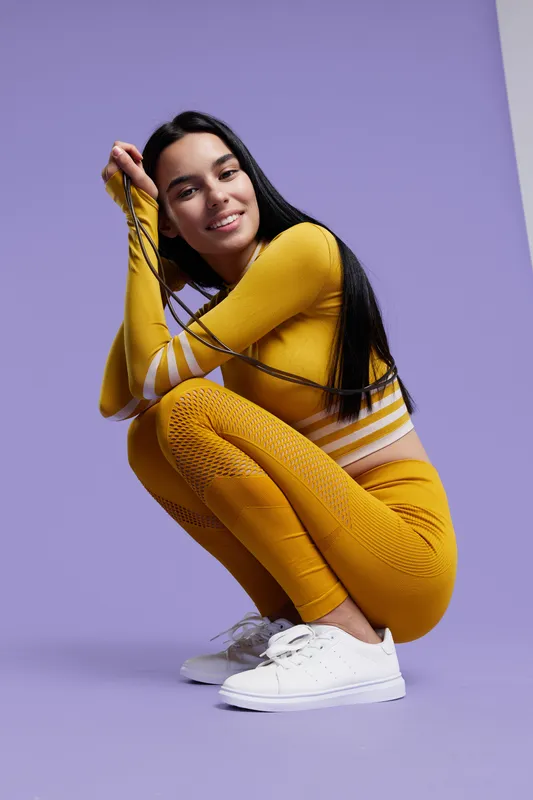 Woman sport fitness in yellow activewear crouching on purple background, smiling with long black hair and white sneakers, healthy lifestyle and workout concept.