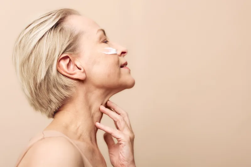 Mature woman with short blonde hair applies white facial cream on cheek, eyes closed, and touches neck gently. Concept of skincare, aging skin care, wellness, and self-care routine.