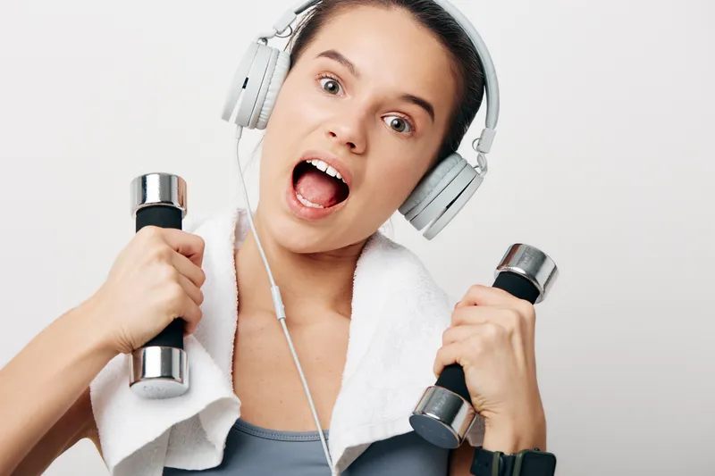 Young woman fitness athlete wearing headphones holds chrome dumbbells with white towel draped over shoulders, energetic expression, studio white background, sporty workout portrait.
