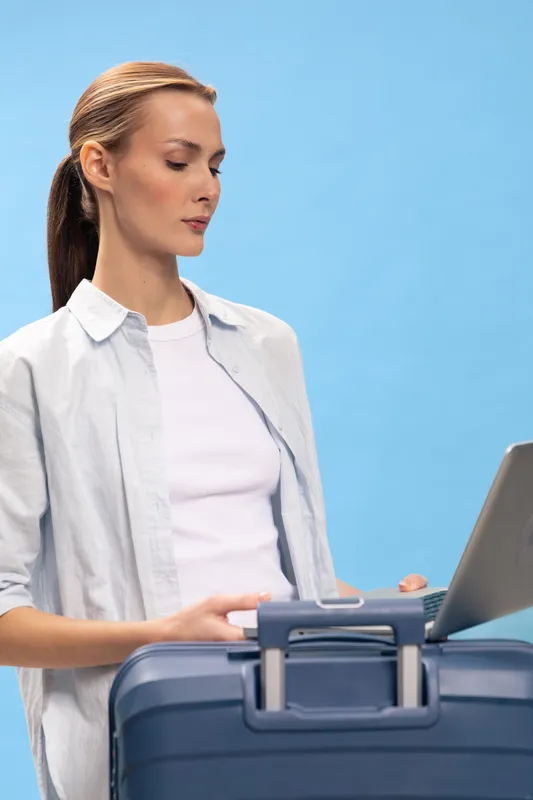 Young woman with suitcase and laptop stands against blue background, focused on screen. Concept of travel planning, business trip, and modern lifestyle.
