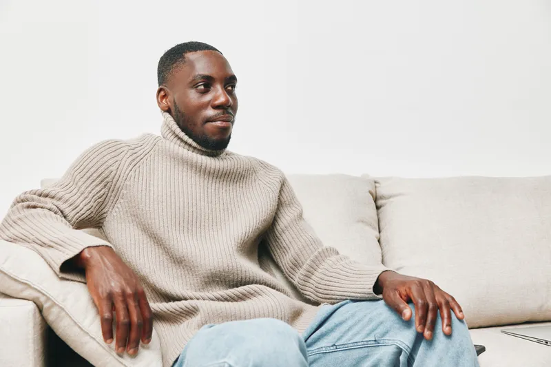 Relaxed young man in a cozy beige turtleneck sweater sitting comfortably on a beige couch, exuding a sense of tranquility and warmth in a minimalist setting