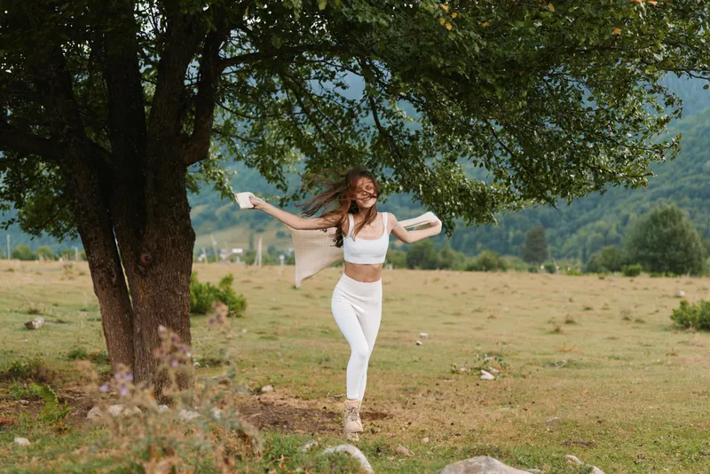 A woman dances freely in a sunlit meadow beside a tree, wearing white clothing, embodying carefree outdoor living, movement, and joyful expression.