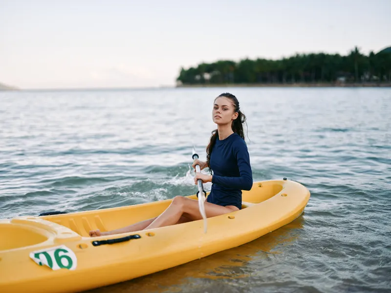 A young paddler sits in a bright yellow kayak on calm coastal water, gripping a paddle as the shoreline stretches under a clear sky.