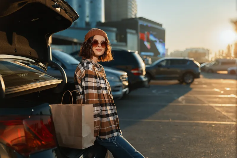 Woman shopping by car in a parking lot at golden hour, sunglasses on, holding a bag while smiling. Urban candid lifestyle shot with authenticity and emotional storytelling.