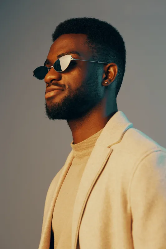 Stylish young African American man in sunglasses and a beige sweater with a confident smile against a soft gradient background showcasing modern fashion and urban aesthetics