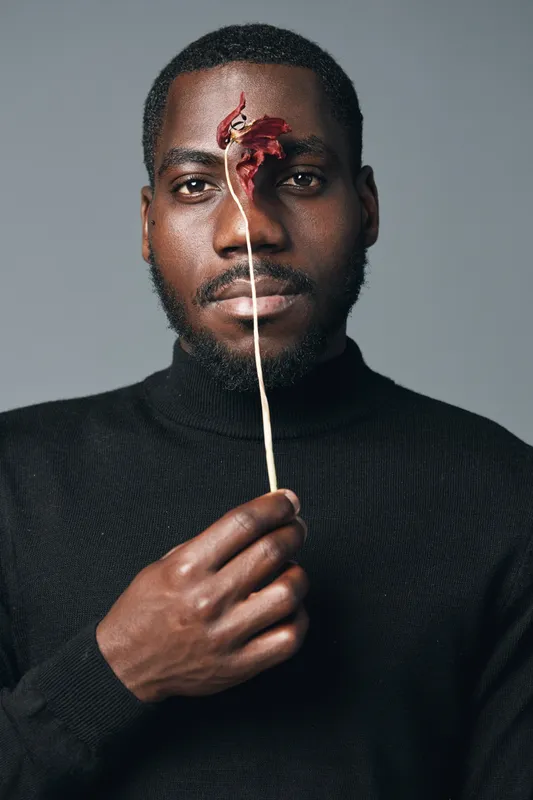 African man with dark skin wearing black turtleneck holding dried red flower in front of face in creative conceptual visual style with minimal gray background and serious expression.