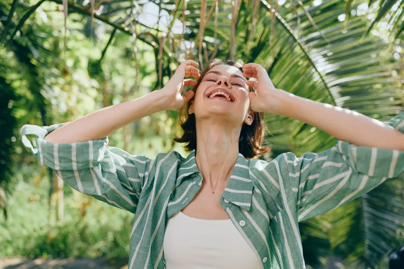 Young woman outdoors in tropical greenery, smiling with hands in hair, casual striped shirt, sunlit moment of joy and freedom.