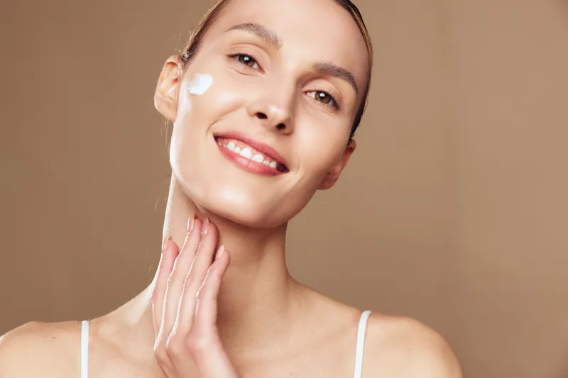 Smiling woman with clear skin applying white face cream on cheek and neck. Close-up on beige background. Concept of skincare, beauty routine, wellness, and self-care with soft lighting.