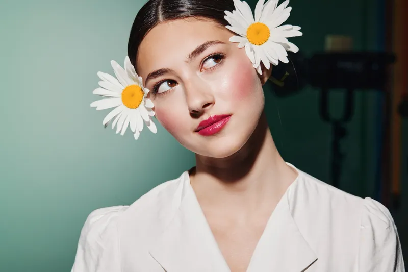 Young woman in white outfit with clear skin and daisies in her hair poses looking upwards. Beauty creative shot focusing on fresh nature-inspired fashion and floral detail.