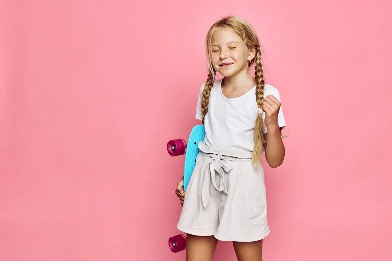 Happy young girl with long braided hair holds a blue skateboard and clenches her fist with eyes closed. Pink background with bright, cheerful studio lighting. Concept of childhood fun and sport.