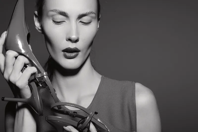 Young Caucasian woman with pale skin holds shiny high heel shoes near her face, eyes closed. Concept of fashion and style.