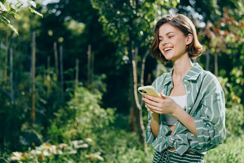 A cheerful woman stands outdoors in a sunny park, smiling while looking at her phone, wearing a striped shirt and casual jacket, enjoying a relaxed moment.