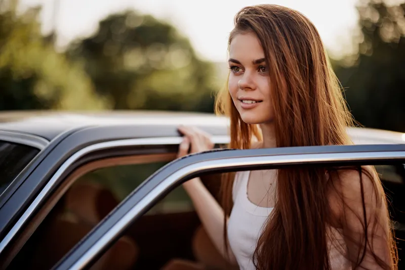 Young woman with long hair smiling next to a vintage car in nature during golden hour, showcasing a joyful, relaxed vibe and warm color palette.