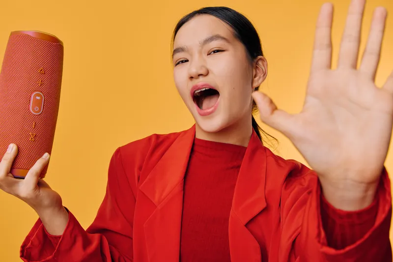 Young woman in red jacket holding wireless speaker singing or shouting on yellow background with hand raised for expression emotion and lifestyle music enjoyment.
