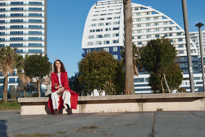 A woman in a red coat sits on a concrete bench in an urban plaza, with modern buildings behind. A calm, stylish moment captured in bright daylight.