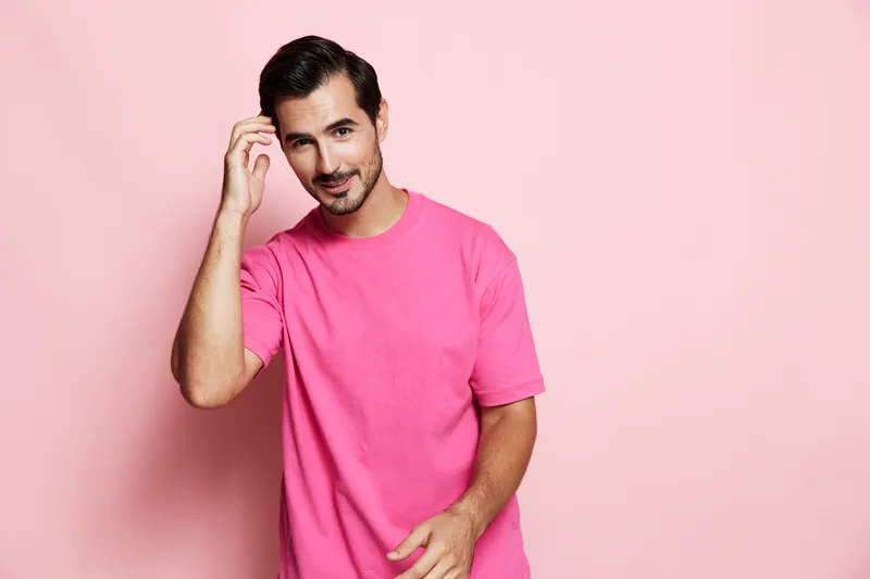 Young smiling man wearing bright pink t-shirt looks at the camera, expressing a cheerful emotion, with a friendly gesture against a soft pastel pink background.