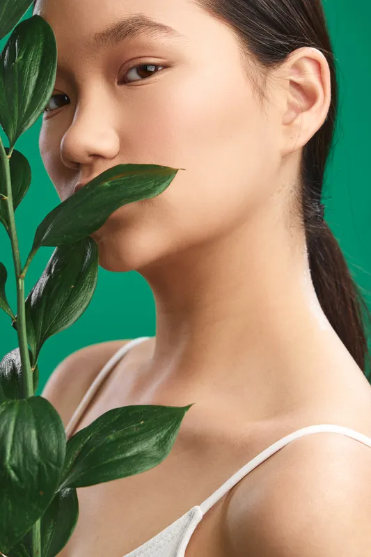 Asian woman gazes softly at camera through leafy plant foreground, highlighting natural beauty and calm aesthetics in a fresh studio setting with a vibrant green backdrop.