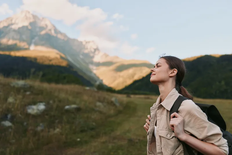 Young woman with a backpack walks through a mountain valley, enjoying the fresh air and warm sunlight.