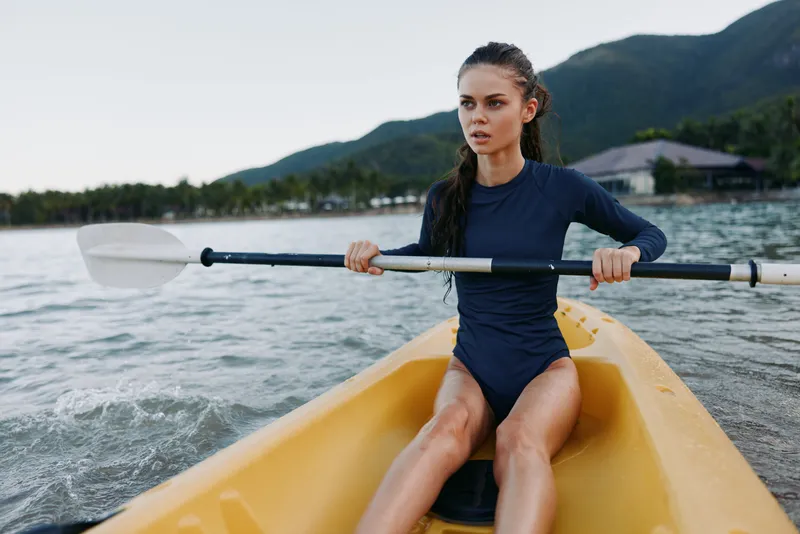 Athletic woman paddling a yellow kayak on calm water, wearing a dark long sleeve swimsuit, focused expression as the oar slices through the tranquil lake.