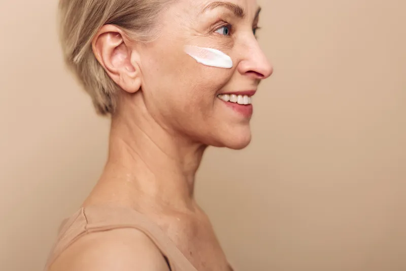 Smiling mature woman with short blonde hair applies white face cream on cheek in beige studio. Concept of skincare, antiaging, natural beauty, wellness, and selfcare for older skin.