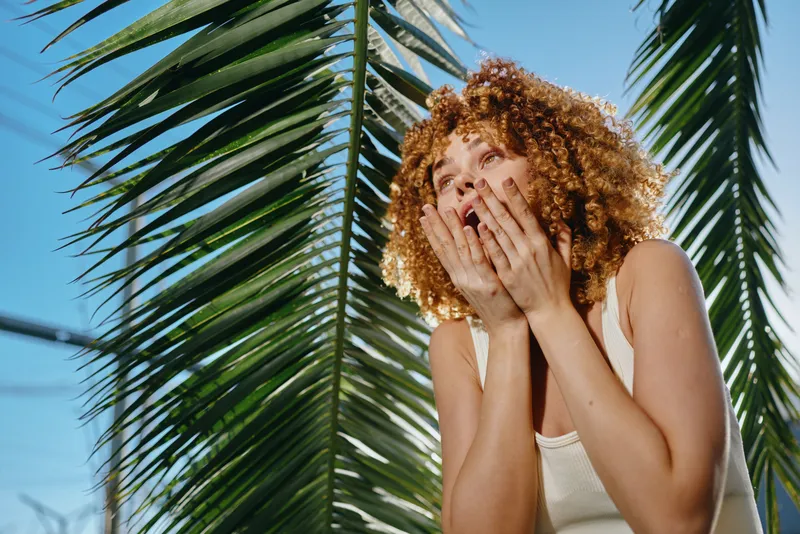 curious woman with curly hair wearing white tank top outdoors under bright sun and green palm leaves showing surprised expression in summer day