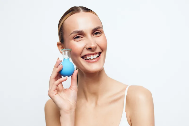Young smiling woman with blonde hair holds a small blue bottle near her face against a white background in a bright studio setting.