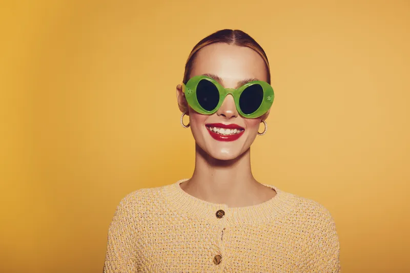 Smiling woman wearing round green sunglasses and yellow sweater poses against yellow background. Concept of fashion, summer style, and cheerful mood with bright colors and accessories.
