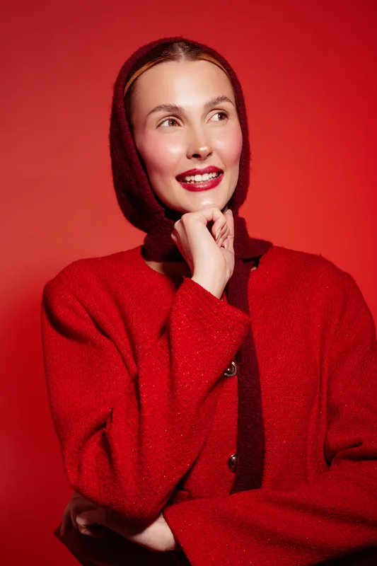 Smiling woman in red wool coat and matching headscarf poses against red background. She looks away with hand near chin, creating a warm and stylish portrait with vibrant colors.