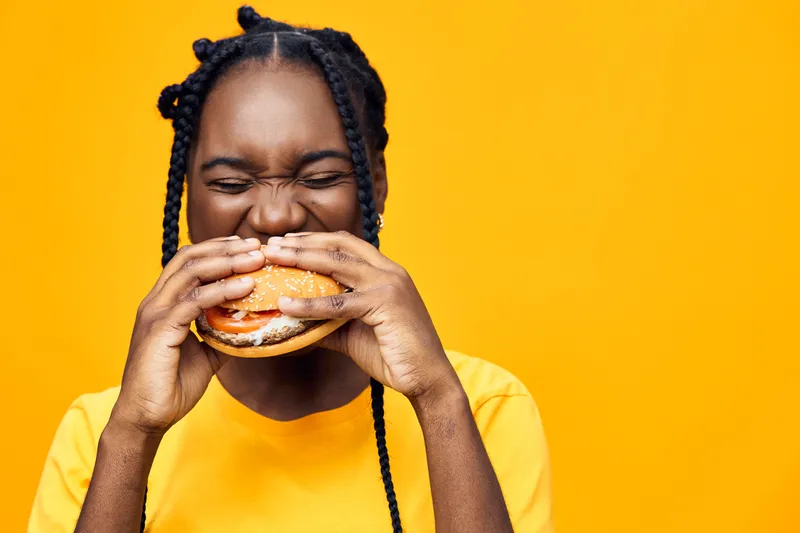 Happy young woman enjoying a delicious burger, smiling brightly against a vibrant yellow background, showcasing food, joy, and casual eating style