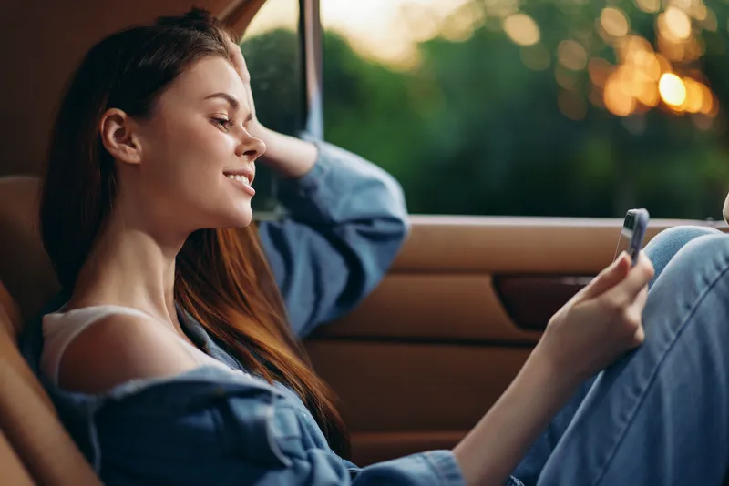 Young woman enjoying a moment of relaxation inside a car, wearing casual denim clothing, smiling while using her phone against a soft sunset backdrop.
