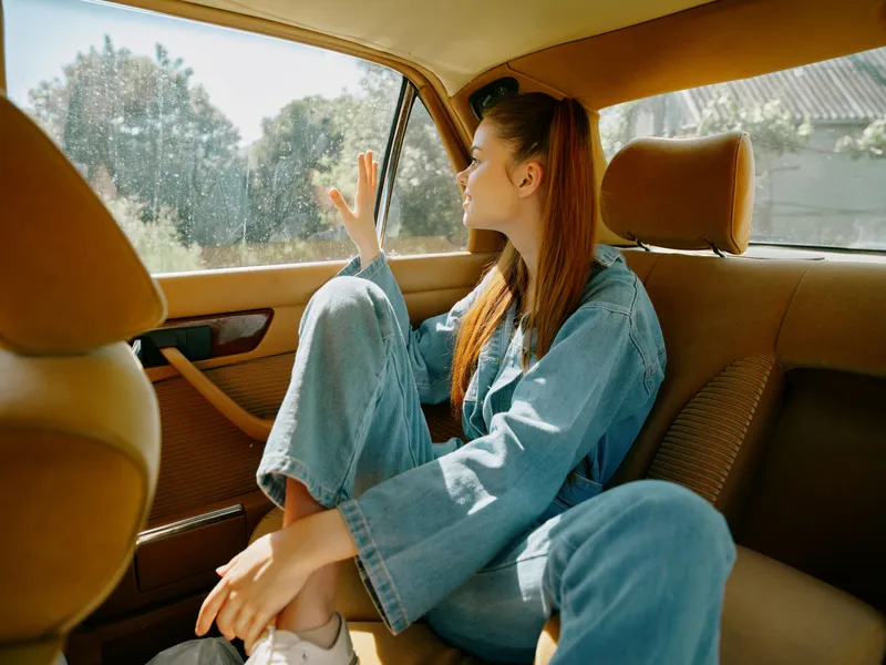 Young woman in denim outfit sitting in a car, looking out the window with a thoughtful expression, reflecting on her journey, in a bright and sunny ambiance.